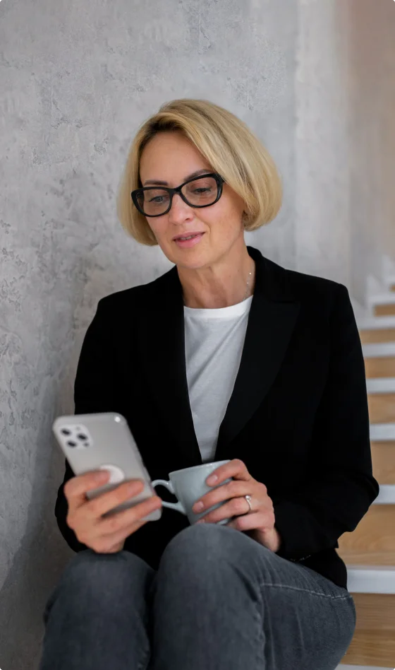 En kvinne med briller sitter og scroller gjennom en sosial medie-feed på iPhonen sin mens hun drikker kaffe