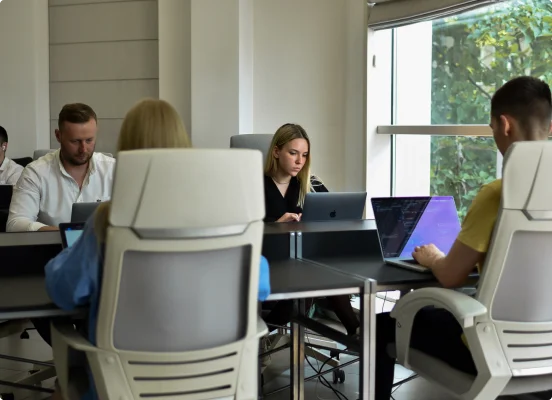 Fünf IT-Experten, zwei Frauen und drei Männer, arbeiten mit ihren Laptops und sitzen auf milchweißen Stühlen hinter ihren Schreibtischen