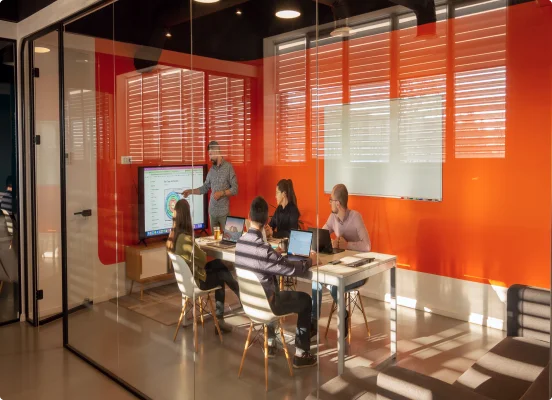 Mehrere IT-Beratungs- und Strategieexperten versammeln sich in einem roten Raum mit geöffneten Laptops und diskutieren Pläne unter Verwendung einer Tafel
